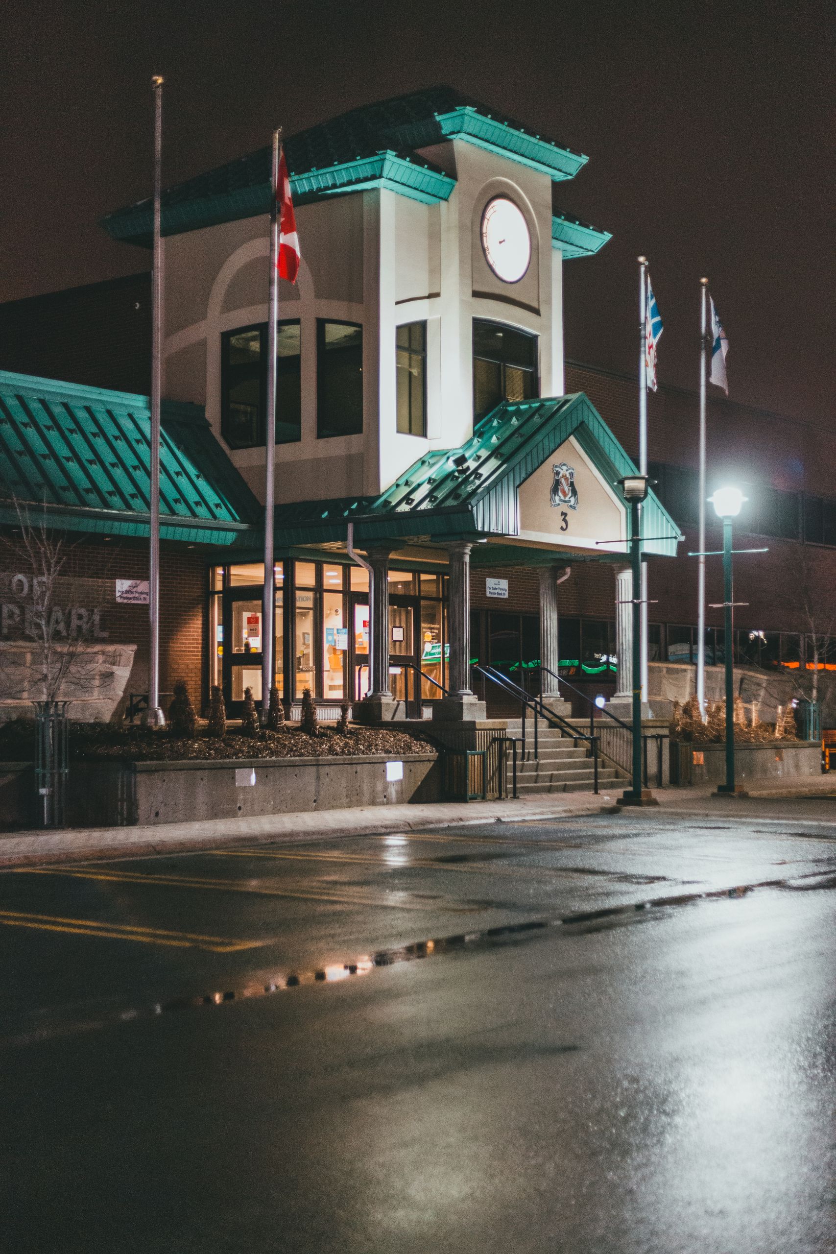 Facade of building with glass walls and clock tower near flags on poles and asphalt in nighttime