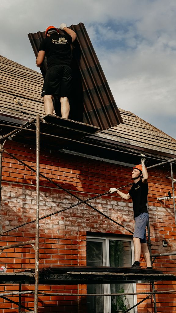 Two builders on scaffolding install panels on a brick building roof.