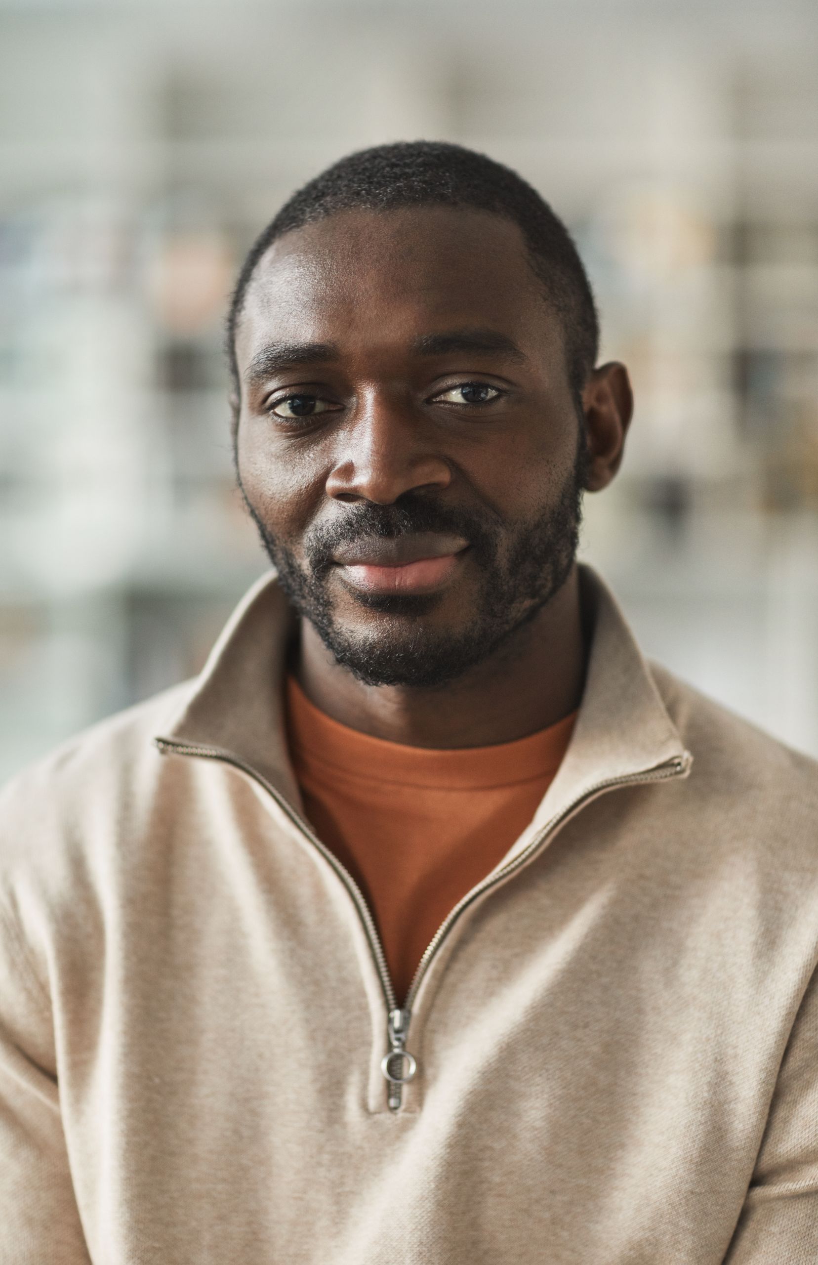 Portrait of a smiling man with facial hair in a beige jacket indoors.